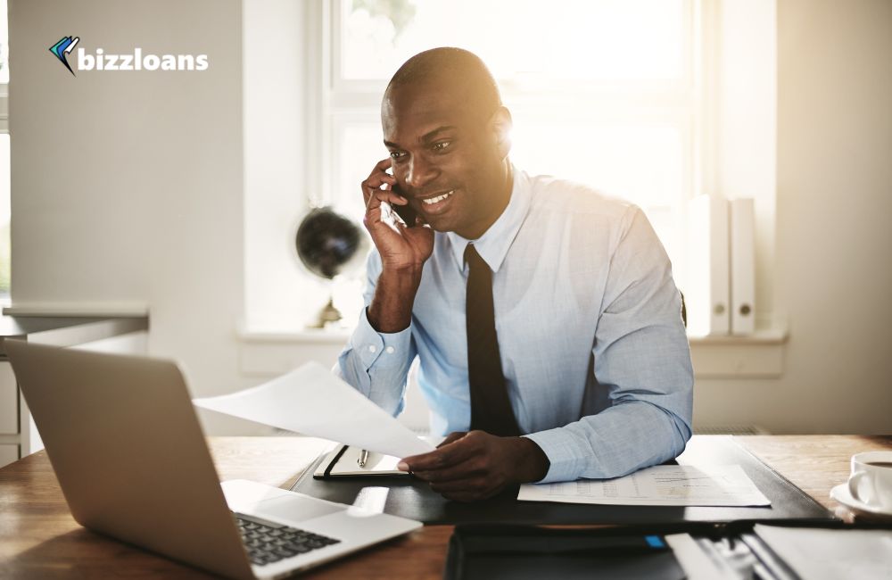 male broker talking to a client on the phone while holding a paper in front of laptop